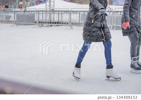 People ice skating outdoors during snowfall, dressed in warm winter clothes. Concept of winter activity, cold season lifestyle, active outdoor fun. Winter background, copy space People ice skating outdoors during snowfall, dressed in warm winter clothes. Concept of winter activity, cold season lifestyle, active outdoor fun. Winter background, copy space 134309152