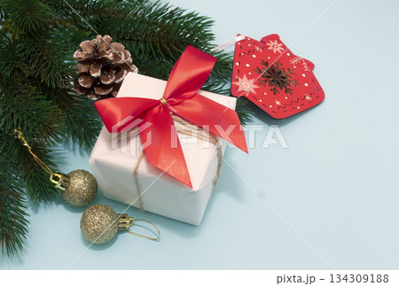 White gift box with red ribbon, gold ornaments, pinecone and mitten tag on light blue background, festive and cheerful White gift box with red ribbon, gold ornaments, pinecone and mitten tag on light blue background, festive and cheerful 134309188