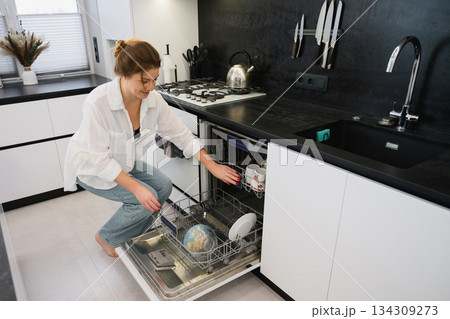 Woman loading dishwasher while crouching in modern kitchen. 134309273