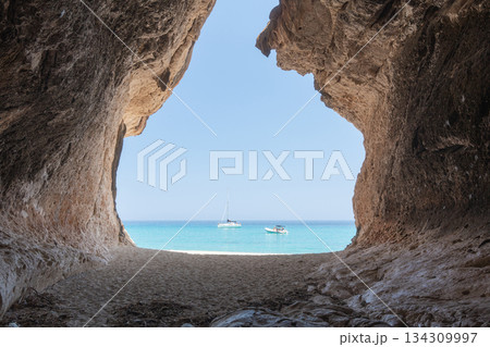 A dramatic dark cave frames a bright seascape with anchored boats in the distance. The strong contrast between shadowed rocks and sunlit turquoise water creates a striking coastal composition 134309997
