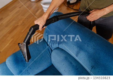 High-angle view of cleaning service worker using washing vacuum cleaner to remove dirt and stains from blue sofa in living room, ensuring clean and healthy environment. Concept of cleanliness. 134310823