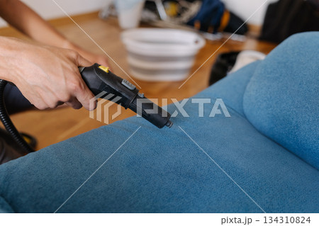 Housekeeper using steam cleaner to remove dirt and stains from blue sofa in living room, ensuring hygiene and freshness, close-up. Concept of maintenance modern home appliance. 134310824