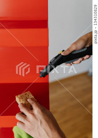 Vertical cropped shot of man using modern appliance to housekeeping, cleaning red shelf using steam cleaner and sponge, ensuring hygiene and spotless surface, closeup. Concept of hygiene, cleanliness. 134310829