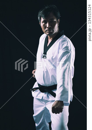 Martial artist in Taekwondo uniform and black belt standing confidently in ready stance against dark background, representing discipline, strength, and combat readiness Martial artist in Taekwondo uniform and black belt standing confidently in ready stance against dark background, representing discipline, strength, and combat readiness 134315124