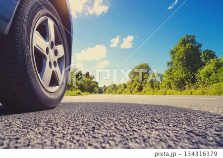 Low Angle Close-Up of Sunlit Car Tire on Asphalt Road Surrounded by Lush Greenery Low Angle Close-Up of Sunlit Car Tire on Asphalt Road Surrounded by Lush Greenery 134316379