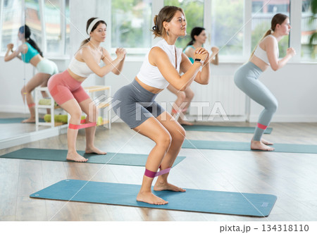 Young woman during Pilates strength training performs squats with elastic band on legs and shins 134318110