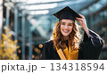 Young graduate female smiling and touching graduation cap while holding diploma. 134318594