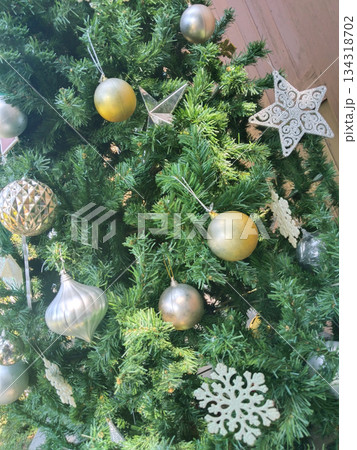 Decorated Christmas tree with ornaments and decorations. The ornaments include golden spheres, silver bulb-shaped objects, snowflake-shaped objects, and a star-shaped ornament Decorated Christmas tree with ornaments and decorations. The ornaments include golden spheres, silver bulb-shaped objects, snowflake-shaped objects, and a star-shaped ornament 134318702