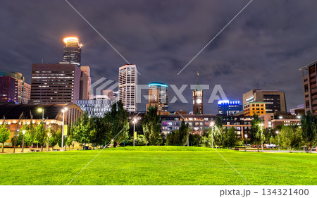 Night view of downtown Minneapolis skyline from The Commons park. Illuminated skyscrapers and historic Armory rise beyond the green lawn under a cloudy sky 134321400