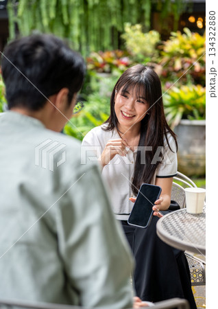 Pretty asian woman holding and showing phone to man while sitting on chair at table in cafe's garden Pretty asian woman holding and showing phone to man while sitting on chair at table in cafe's garden 134322880