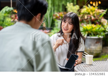Pretty asian woman holding or showing phone to man while sitting on chair at table in cafe's garden 134322881