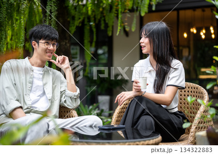 Pretty asian woman holding coffee talking with a glasses man while sitting at table in cafe's garden 134322883