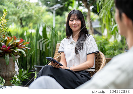 Pretty asian woman holding phone looking at a man friend while sitting on woven chair in cafe garden 134322888