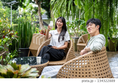 Pretty asian woman holding phone pointing for man friend while sitting on woven chair in cafe garden 134322889