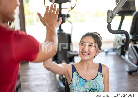 Fitness trainer coach clapping hands high five with asian woman client on exercise equipment in gym. 134324061