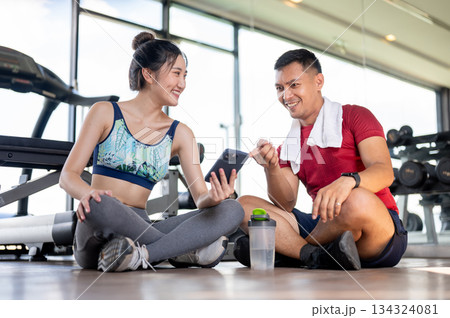 Asian woman client showing phone to fitness coach trainer man sits on floor after exercising in gym. 134324081