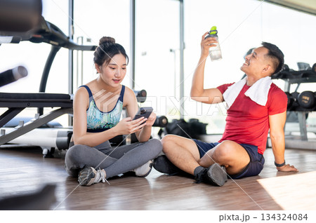 Woman client looking at phone and fitness gym trainer drinking water sits on floor after exercising. Woman client looking at phone and fitness gym trainer drinking water sits on floor after exercising. 134324084