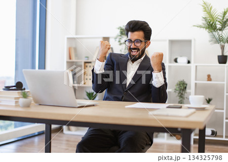 An excited businessman celebrates a win in his office, looking at his laptop screen An excited businessman celebrates a win in his office, looking at his laptop screen 134325798
