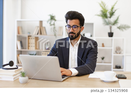 An Arabian businessman wearing glasses is working on a laptop in a modern office setting 134325908
