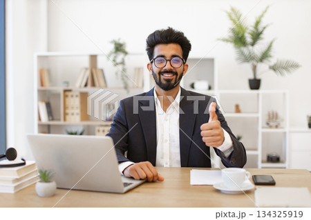 An Indian businessman in a suit smiles and gives a thumbs up while working on his laptop 134325919