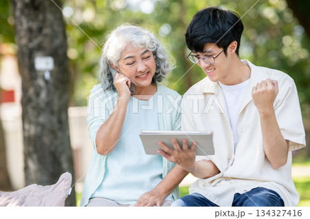 Old woman grandmother looking at tablet and young man grandson making a fist while sitting in park. 134327416