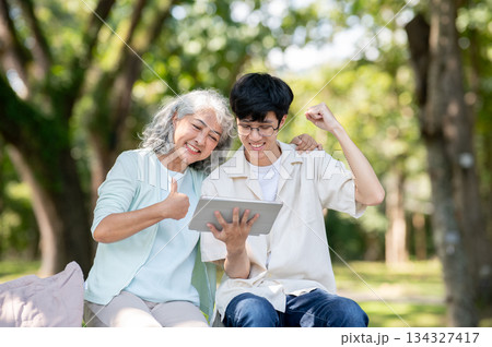 Thumb up old woman grandmother looking at tablet and young man grandson raising a fist sits in park. Thumb up old woman grandmother looking at tablet and young man grandson raising a fist sits in park. 134327417