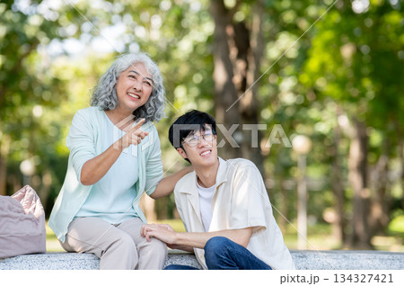 Old woman grandmother pointing aside young man grandson while sitting on stone bench in garden park. Old woman grandmother pointing aside young man grandson while sitting on stone bench in garden park. 134327421
