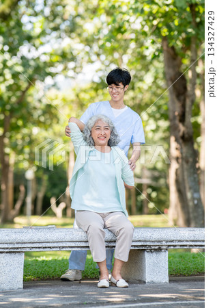 Glasses man caregiver nurse helping old woman granny patient exercise while sitting on bench in park Glasses man caregiver nurse helping old woman granny patient exercise while sitting on bench in park 134327429