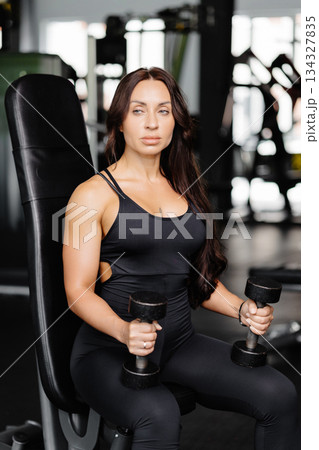 Young fit woman practicing dumbbell press while sitting on a bench. Slim female in sportswear exercises with weights in a gym Young fit woman practicing dumbbell press while sitting on a bench. Slim female in sportswear exercises with weights in a gym 134327835