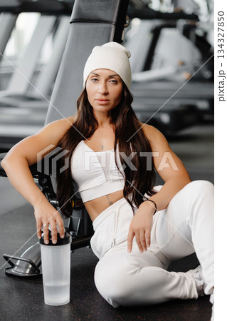 Strong woman with long hair is sitting on the floor in the gym on the background of the simulators. She is wearing white sportswear with white sneakers. 134327850