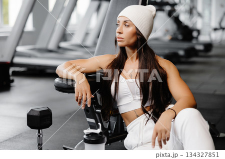 Strong woman with long hair is sitting on the floor in the gym on the background of the simulators. Strong woman with long hair is sitting on the floor in the gym on the background of the simulators. 134327851
