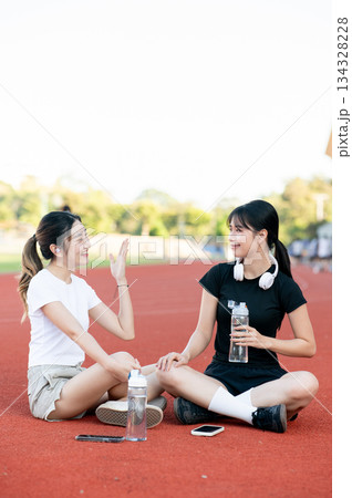 Asian woman holding water talking to a friend as sitting on running track ground in sports stadium. 134328228