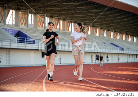 Happy asian woman friend running jogging together on racetrack exercising outdoors in sports stadium 134328233