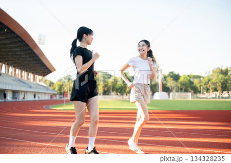 Asian woman and friend talking while running or jogging on racetrack in sports stadium on sunny day. Asian woman and friend talking while running or jogging on racetrack in sports stadium on sunny day. 134328235