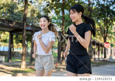 Pretty asian woman and friend smiling while running or jogging on concrete road exercising in a park Pretty asian woman and friend smiling while running or jogging on concrete road exercising in a park 134328251