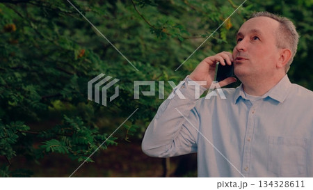 casual attire, casual blue shirt, listening intently happily., smiling caller in nature, standing outside amid lush natural surroundings., standing by leafy branches nearby., happy middle-aged man 134328611