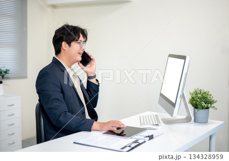 Smiling glasses asian businessman phone calling coworker client at computer table in company office Smiling glasses asian businessman phone calling coworker client at computer table in company office 134328959