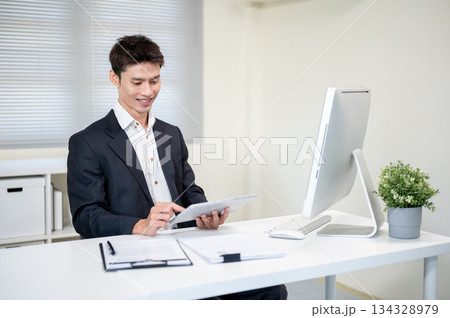 Smiling asian businessman holding and looking at tablet aside computer on table in company's office. Smiling asian businessman holding and looking at tablet aside computer on table in company's office. 134328979