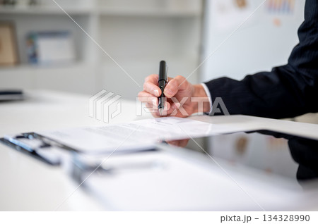 Close up businessman's hand holding pen writing in document on clipboard at working table in office. Close up businessman's hand holding pen writing in document on clipboard at working table in office. 134328990