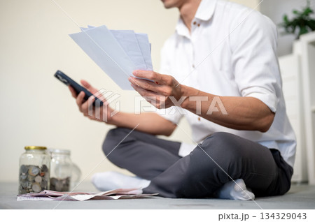Close up of office worker man holding phone and bills as sitting on living room floor with coin jars Close up of office worker man holding phone and bills as sitting on living room floor with coin jars 134329043