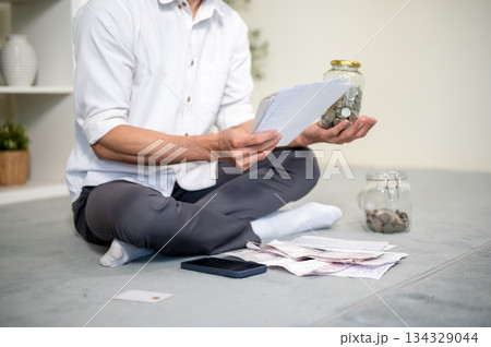 Close up of office worker man holding coin jar looking at bills while sitting on living room floor. Close up of office worker man holding coin jar looking at bills while sitting on living room floor. 134329044