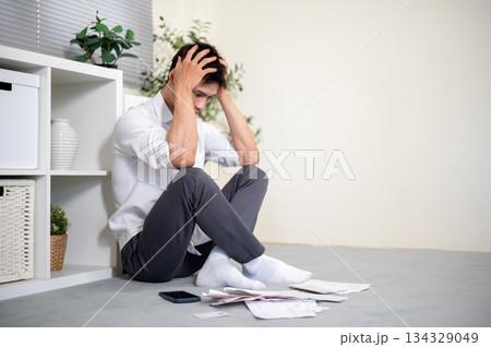 Frustrated asian man office worker holding head looking at bills as sitting on the living room floor 134329049