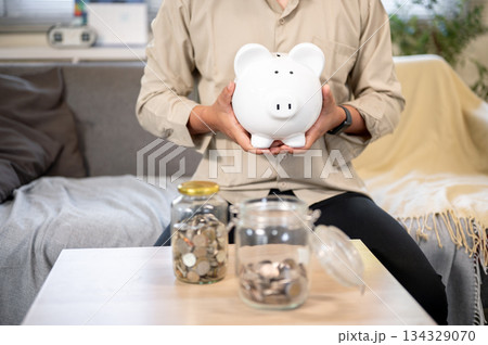 Close up office worker man holding a piggy bank aside coin glass jars sitting on sofa in living room 134329070