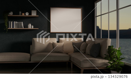Blank frame and wooden shelf over sofa couch in dim living room with sunrise outside glass wall window. 134329076