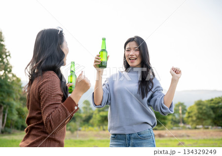 Asian woman and friend holding drink bottle dancing in green grass field with mountain view and sky. Asian woman and friend holding drink bottle dancing in green grass field with mountain view and sky. 134329249