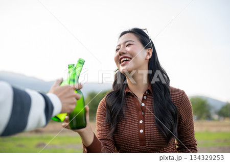 Pretty asian woman clinking drink bottle with friend in grass field with mountain view and clear sky Pretty asian woman clinking drink bottle with friend in grass field with mountain view and clear sky 134329253