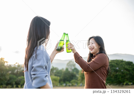 Asian woman and friend holding alcohol drink and clinking bottle in field under sky and sunlight. Asian woman and friend holding alcohol drink and clinking bottle in field under sky and sunlight. 134329261