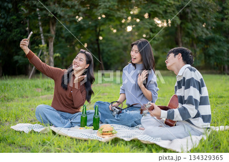 Asian friends woman and man taking picture on phone while sitting on picnic mat in green grass field Asian friends woman and man taking picture on phone while sitting on picnic mat in green grass field 134329365