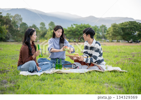 Asian woman drinking and eating burger and man friend with guitar on picnic mat in green grass field 134329369