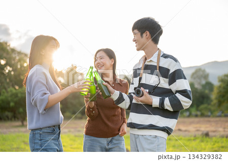 Asian friends woman and a man clinking drink bottles in grass field with mountain view and sunlight. Asian friends woman and a man clinking drink bottles in grass field with mountain view and sunlight. 134329382